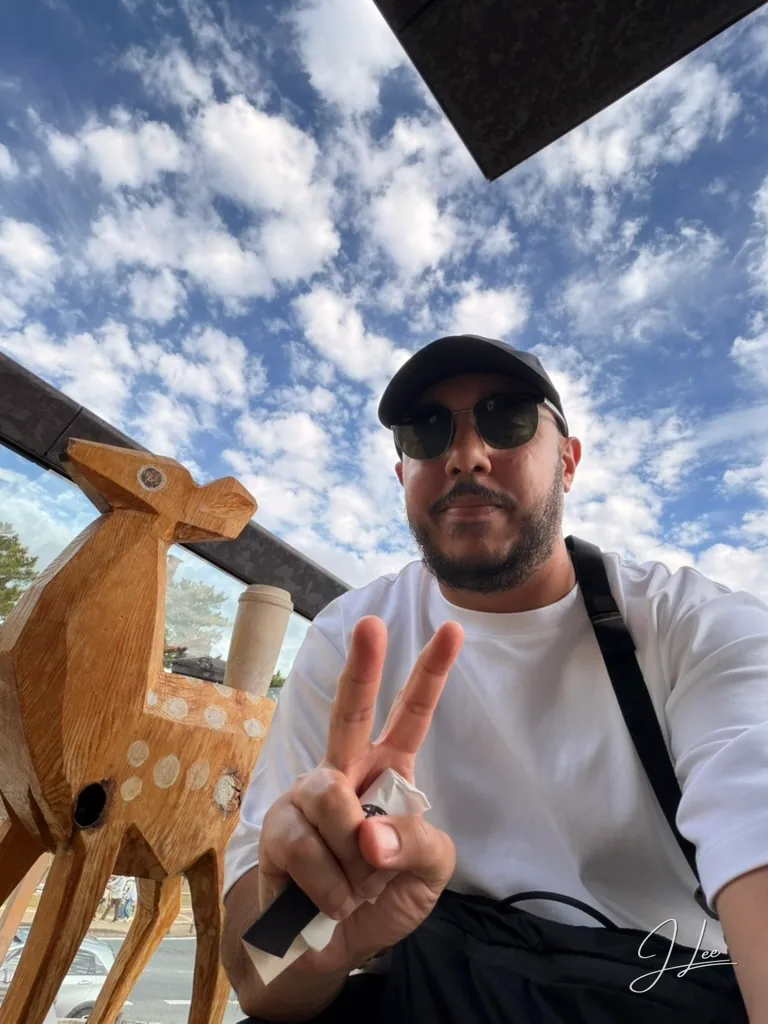 Jordan Lee outdoors beneath a bright blue sky, wearing sunglasses and making a peace sign beside a wooden deer sculpture.