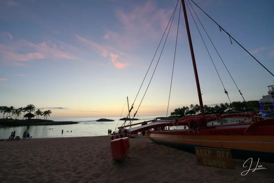 A red outrigger canoe rests on the sand beside a calm bay under a pastel sky.