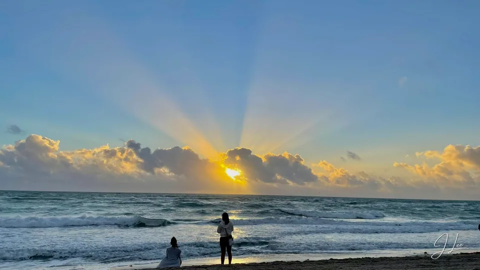 Sunbeams break through clouds over ocean waves, with two people at the shoreline.