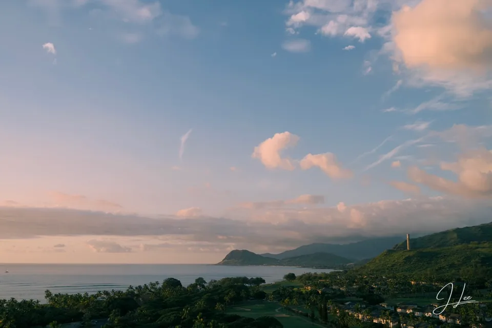 Soft pink clouds above a coastal bay, green hills, and palm-lined grounds.