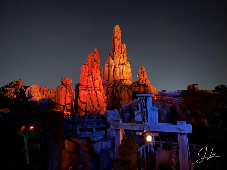 Orange-lit rock spires above blue-lit wooden structures at night.