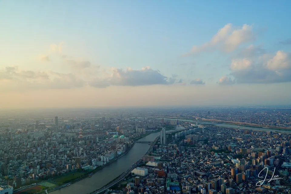 A broad cityscape and curving river seen from above beneath a hazy, cloud-filled sky.
